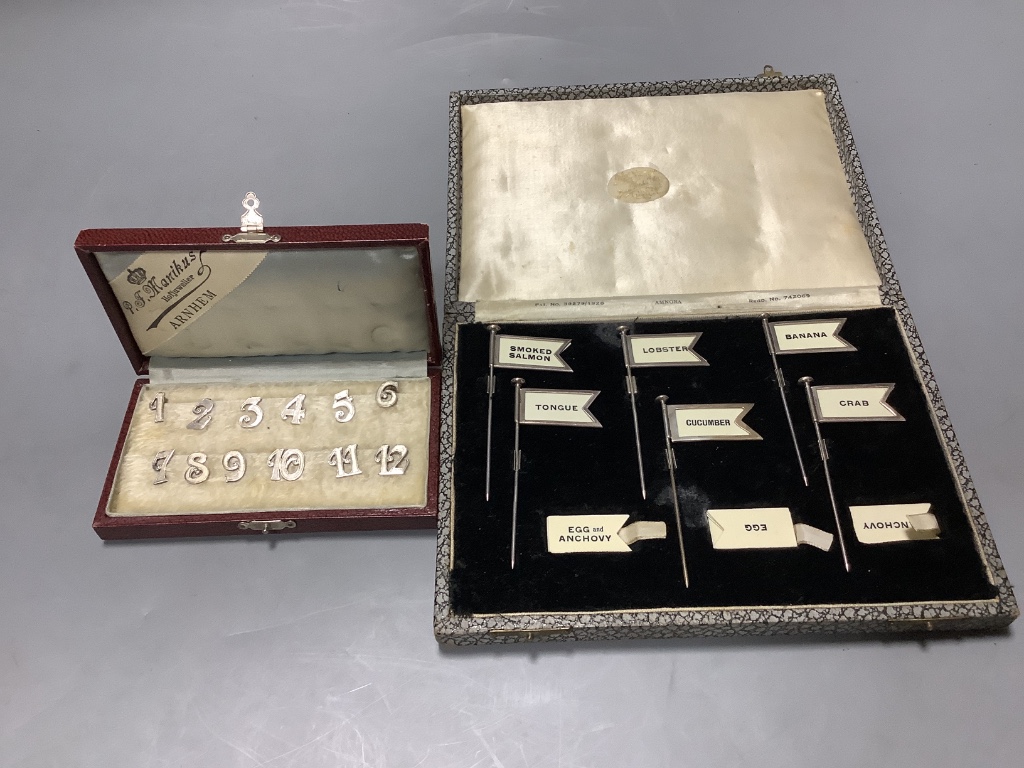 A cased set of twelve Dutch white metal number place settings, 12mm and a cased set of six silver sandwich flags sticks, height 8cm.